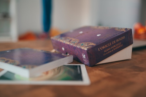 A deck of oracle cards is prominently placed on a wooden table. The packaging is purple with intricate gold and light patterns, and the name 'L'Oracle De Rumi' by Alana Fairchild with illustrations by Rassouli is visible. Beside the box, some cards are partially visible, slightly out of focus, adding a mystical and calm ambiance to the scene.