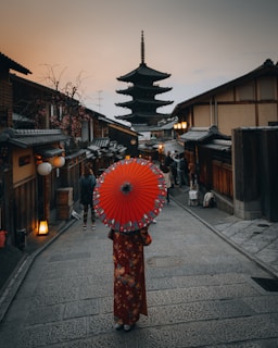 A person wearing a traditional kimono holds a vibrant red parasol, walking down a narrow, paved street lined with wooden buildings. Overhead, the sky is overcast with a pagoda rising in the distance, while warm street lamps begin to glow.
