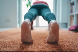 A person is in a downward-facing position on an orange carpet, wearing dark leggings. The focus is on their feet and lower legs as they perform a yoga pose.