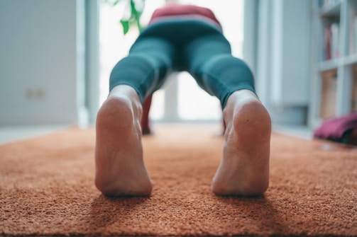 A person is in a downward-facing position on an orange carpet, wearing dark leggings. The focus is on their feet and lower legs as they perform a yoga pose.