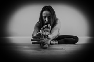 A person performs a seated forward bend stretch on a wooden floor, with focus on holding their feet and extending their body forward. The background is a plain wall, and the image is captured in black and white, creating a stark contrast and a moody atmosphere.