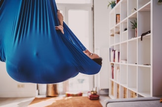A person is wrapped and suspended in a blue fabric hammock, positioned upside down. The setting is a cozy room with a soft carpet, a shelving unit holding books and plants, and warm, natural lighting coming through large windows.