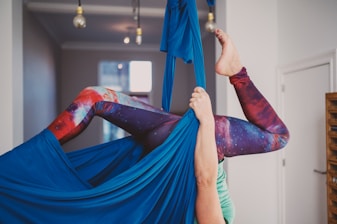 A person is performing aerial yoga in an indoor space. They are suspended in fabric, with their legs bent and elevated. The person is wearing colorful leggings with a galaxy pattern, and their hands are gripping the blue aerial silk. The room has a modern look with several light bulbs hanging from the ceiling and a window in the background.