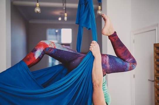 A person is performing aerial yoga in an indoor space. They are suspended in fabric, with their legs bent and elevated. The person is wearing colorful leggings with a galaxy pattern, and their hands are gripping the blue aerial silk. The room has a modern look with several light bulbs hanging from the ceiling and a window in the background.