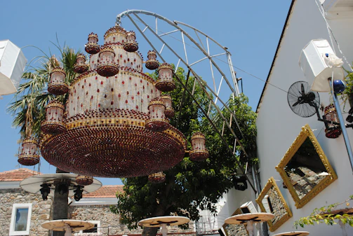 An ornate chandelier made of intricate glass and metalwork hangs outdoors. The chandelier features multiple tiers and numerous decorative lamps. Behind the chandelier, a tree with lush green leaves is visible, alongside a white building with several square, gold-framed mirrors on the wall. Patio tables with umbrellas are positioned beneath the chandelier, and a fan is mounted on the wall.