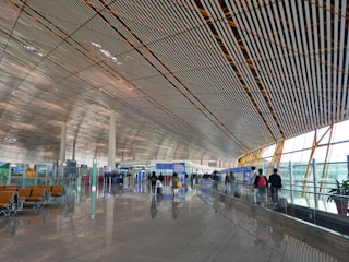 A spacious and modern airport terminal with a high, patterned ceiling and large windows allowing natural light to illuminate the area. People are walking along the concourse with their luggage, and there are empty seats available for waiting passengers. Signs and information boards are visible, providing travel information.