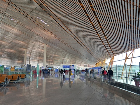 A spacious and modern airport terminal with a high, patterned ceiling and large windows allowing natural light to illuminate the area. People are walking along the concourse with their luggage, and there are empty seats available for waiting passengers. Signs and information boards are visible, providing travel information.