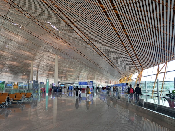 A spacious and modern airport terminal with a high, patterned ceiling and large windows allowing natural light to illuminate the area. People are walking along the concourse with their luggage, and there are empty seats available for waiting passengers. Signs and information boards are visible, providing travel information.