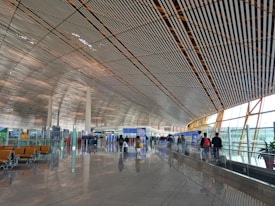 A spacious and modern airport terminal with a high, patterned ceiling and large windows allowing natural light to illuminate the area. People are walking along the concourse with their luggage, and there are empty seats available for waiting passengers. Signs and information boards are visible, providing travel information.