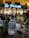 A bottle of mineral water labeled 'aqua noble' sits on a table next to a tall sugar tin and a patterned glass under low light. In the background, a softly lit café named 'Le Pristato' is visible with patrons sitting inside and outside, creating a cozy and lively ambiance.