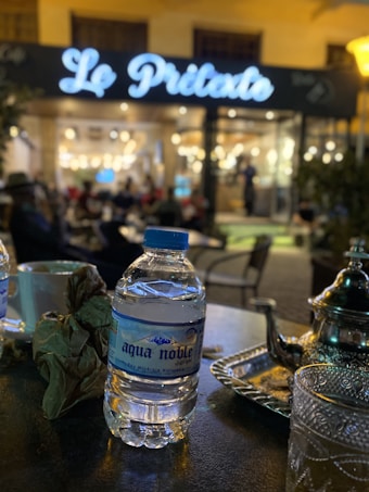 A bottle of mineral water labeled 'aqua noble' sits on a table next to a tall sugar tin and a patterned glass under low light. In the background, a softly lit caf&eacute; named 'Le Pristato' is visible with patrons sitting inside and outside, creating a cozy and lively ambiance.