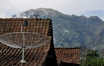 A satellite dish is mounted on a roof with a reddish-brown tiled pattern against a backdrop of a mountainous landscape. The sky is partly cloudy, and a bird is perched on the edge of the roof.