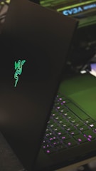 A sleek black laptop with a glowing green logo on its lid, partially open to reveal a keyboard with illuminated keys in shades of purple. The setting appears to be a dark environment, highlighting the luminous elements of the device.