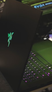 A sleek black laptop with a glowing green logo on its lid, partially open to reveal a keyboard with illuminated keys in shades of purple. The setting appears to be a dark environment, highlighting the luminous elements of the device.