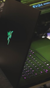 A sleek black laptop with a glowing green logo on its lid, partially open to reveal a keyboard with illuminated keys in shades of purple. The setting appears to be a dark environment, highlighting the luminous elements of the device.