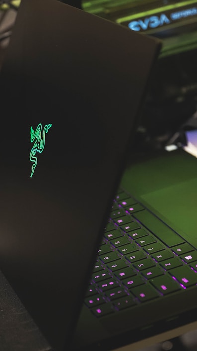 A sleek black laptop with a glowing green logo on its lid, partially open to reveal a keyboard with illuminated keys in shades of purple. The setting appears to be a dark environment, highlighting the luminous elements of the device.