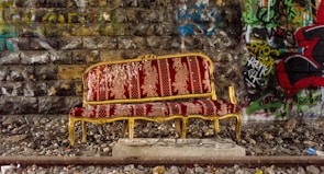 A vintage red upholstered sofa with gold accents sits on a rocky surface in front of a graffiti-covered stone wall. The contrast between the ornate furniture and the gritty urban environment adds an intriguing juxtaposition.