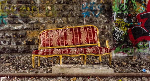 A vintage red upholstered sofa with gold accents sits on a rocky surface in front of a graffiti-covered stone wall. The contrast between the ornate furniture and the gritty urban environment adds an intriguing juxtaposition.