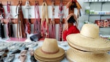 A display of various equestrian gear and accessories is arranged on shelves and hooks. The foreground shows stacks of straw hats and a single hat placed separately. The background features horse bridles, reins, and other leather straps hanging on wooden racks. Shelves in the background contain an assortment of horse care products in bottles and boxes.