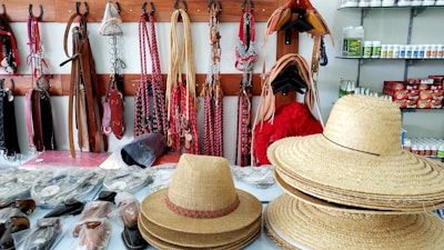 A display of various equestrian gear and accessories is arranged on shelves and hooks. The foreground shows stacks of straw hats and a single hat placed separately. The background features horse bridles, reins, and other leather straps hanging on wooden racks. Shelves in the background contain an assortment of horse care products in bottles and boxes.