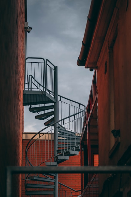 A metal spiral staircase is positioned between two buildings. The staircase has a modern design with a sturdy metal railing. The surrounding buildings have a reddish-brown hue, indicating brick or painted surfaces, and the sky above appears overcast, casting a muted light on the scene.