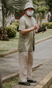 An elderly person stands on a sidewalk, holding a wooden stick and a dark object. They are wearing a light-colored bucket hat, a beige mask, a light green short-sleeved shirt, and cream trousers. The background features palm trees and greenery, suggesting an outdoor park-like setting.