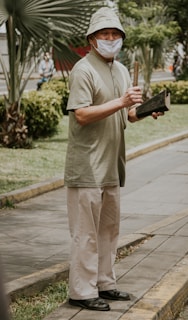 An elderly person stands on a sidewalk, holding a wooden stick and a dark object. They are wearing a light-colored bucket hat, a beige mask, a light green short-sleeved shirt, and cream trousers. The background features palm trees and greenery, suggesting an outdoor park-like setting.