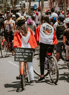 People stand in a crowded outdoor setting, many holding bicycles. Two individuals in the foreground wear capes or flags with a distinctive emblem, suggesting national pride. One holds a sign with a date and message written in Spanish. Various individuals wear helmets and casual clothing, and the environment is urban with buildings and trees visible.