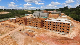 A construction site with multiple unfinished brick apartment buildings surrounded by earth and dirt work in a rural setting. The buildings are in different stages of construction with some showing exposed structural elements. The landscape beyond includes green trees and hills under a partly cloudy sky.
