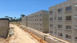 Several unfinished apartment buildings line a dusty construction site. The buildings are rectangular, with windows installed but no finishing or paint applied. Construction materials, such as bricks and concrete pipes, are scattered along the ground. A clear blue sky is visible, with sparse greenery and trees in the background.