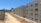Several unfinished apartment buildings line a dusty construction site. The buildings are rectangular, with windows installed but no finishing or paint applied. Construction materials, such as bricks and concrete pipes, are scattered along the ground. A clear blue sky is visible, with sparse greenery and trees in the background.