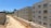Several unfinished apartment buildings line a dusty construction site. The buildings are rectangular, with windows installed but no finishing or paint applied. Construction materials, such as bricks and concrete pipes, are scattered along the ground. A clear blue sky is visible, with sparse greenery and trees in the background.