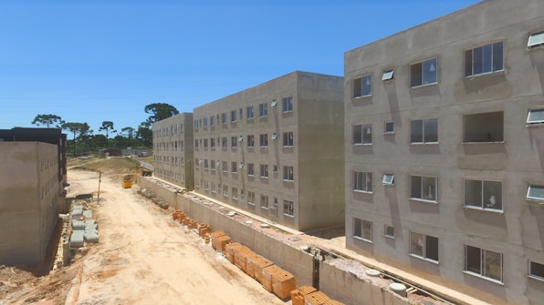Several unfinished apartment buildings line a dusty construction site. The buildings are rectangular, with windows installed but no finishing or paint applied. Construction materials, such as bricks and concrete pipes, are scattered along the ground. A clear blue sky is visible, with sparse greenery and trees in the background.