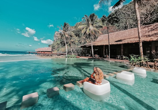 A tropical resort setting with a luxurious poolside scene. A woman with long blonde hair relaxes on a white floating chair in the clear blue water of the infinity pool. The background features palm trees, a thatched-roof structure, and lush greenery. The ocean and sky are visible in the distance, enhancing the tranquil atmosphere.