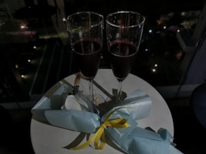 Two tall champagne glasses filled with a dark beverage are placed on a small round table. Next to the glasses is a small bouquet wrapped in light blue paper with a yellow ribbon.