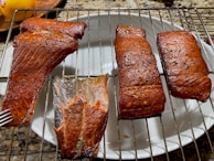 Four pieces of smoked salmon rest on a metal grill rack above a white plate. The salmon pieces are golden brown with a glossy texture and visible seasoning. A fork is placed next to one of the pieces. The background surface has a granite appearance, and part of a yellow apple is visible on the left.