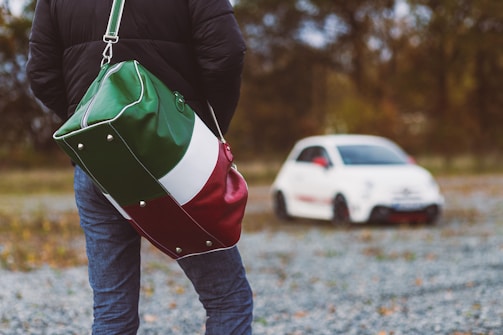 A person wearing a black jacket and jeans stands on a gravel path, facing away with a large duffel bag over their shoulder, designed with a green, white, and red color scheme. In the background, a small white car with red accents is parked at a distance, surrounded by an out-of-focus natural setting with trees and autumn foliage.
