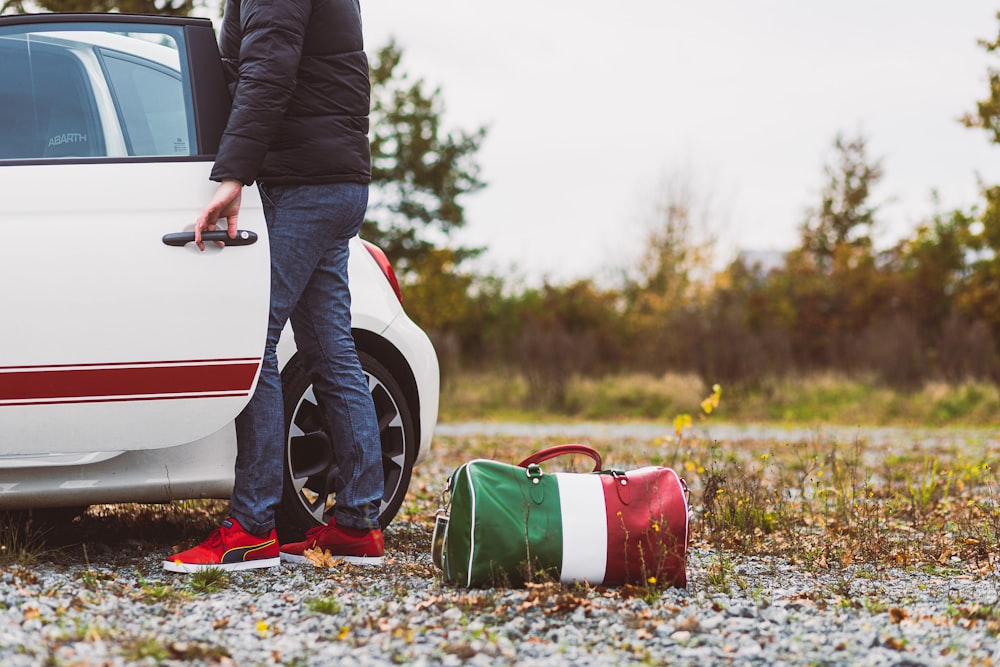 man in black long sleeve shirt and blue denim jeans standing beside white and red car