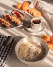 A breakfast setting with a bowl of oatmeal topped with banana slices and sunflower seeds, accompanied by a cup of coffee. On a nearby plate, there are slices of bread with peanut butter and what appears to be a reddish jam, as well as some cookies. Three fresh oranges are also visible on the table, which is covered with a white tablecloth. A checkered napkin with a spoon rests nearby.