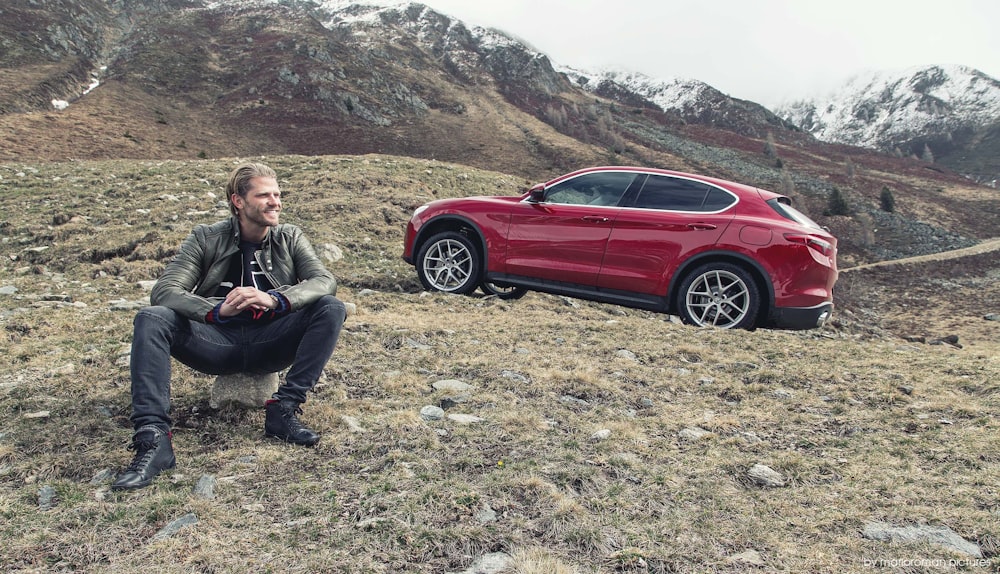 man in black jacket sitting on ground beside red sedan during daytime