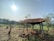 A small rustic shelter with a tiled roof is situated in a rural landscape. It is supported by four pillars and surrounded by dry, bare soil with a few trees and shrubs sparsely scattered around. In the background, there are green, terraced fields extending towards the horizon, with a forested area further back. The sun shines brightly in a clear sky, casting shadows from the shelter and other objects.