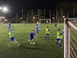A group of young soccer players on a well-lit field at night, engaging intensely in a practice session. Some players are wearing blue jerseys while others are in green bibs, indicating teams or positions. The field is surrounded by a net and illuminated by bright floodlights, with trees visible in the background.