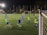 A group of young soccer players on a well-lit field at night, engaging intensely in a practice session. Some players are wearing blue jerseys while others are in green bibs, indicating teams or positions. The field is surrounded by a net and illuminated by bright floodlights, with trees visible in the background.