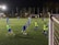 A group of young soccer players on a well-lit field at night, engaging intensely in a practice session. Some players are wearing blue jerseys while others are in green bibs, indicating teams or positions. The field is surrounded by a net and illuminated by bright floodlights, with trees visible in the background.