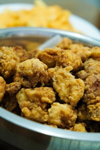 A bowl filled with crispy, golden-brown fried chicken pieces. The chicken appears to be well-seasoned and cooked to a crunchy texture. In the background, there is a blurred dish of food, likely fries or chips.