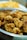 A bowl filled with crispy, golden-brown fried chicken pieces. The chicken appears to be well-seasoned and cooked to a crunchy texture. In the background, there is a blurred dish of food, likely fries or chips.