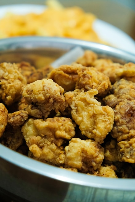 A bowl filled with crispy, golden-brown fried chicken pieces. The chicken appears to be well-seasoned and cooked to a crunchy texture. In the background, there is a blurred dish of food, likely fries or chips.