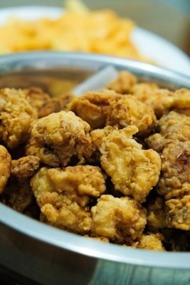A bowl filled with crispy, golden-brown fried chicken pieces. The chicken appears to be well-seasoned and cooked to a crunchy texture. In the background, there is a blurred dish of food, likely fries or chips.