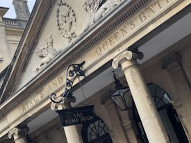 The image portrays a neoclassical building facade with columns and detailed architectural sculptures. Prominent text on the building reads 'KING'S AND QUEEN'S BATH' atop and 'THE PUMP ROOM' on a hanging sign. Ornate lamp fixtures are suspended from the ceiling.