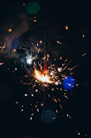 Sparks and bright lights are emanating from a firework against a dark background. Various colored bokeh effects, such as blue and green, appear around the central burst of light.